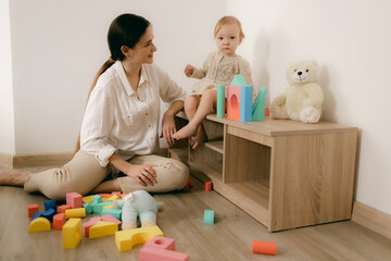 Young happy mother playing game with educational pyramid with cute little child girl in the living room at home. Woman parent spending time with daughter. Family leisure and people concept.