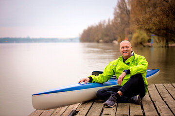 Sporty active senior sitting on the jetty by the river next to his kayak
