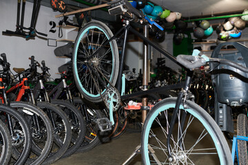 Bicycle standing on repair stand in workshop surrounded by multiple bikes and spare wheels, mechanic tools visible in background, indoor setting focused on maintenance activity