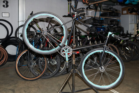 Bicycle with turquoise tires mounted on repair stand undergoing maintenance in workshop, multiple bicycles and spare wheels visible in background, indoor setting, no people present
