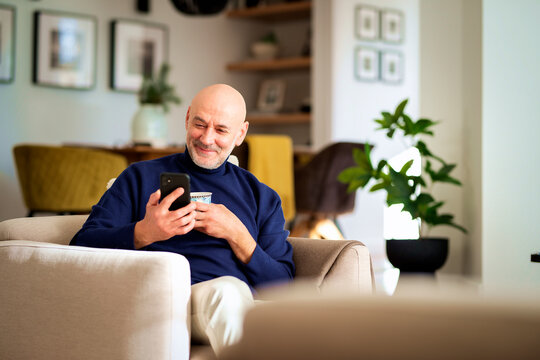 Happy middle-aged man with smartphone relaxing at home and drinking tea - Powered by Adobe