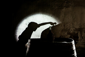 The shadow of a child extends toward a wicker wine bottle resting on an aged oak barrel in dim cellar.