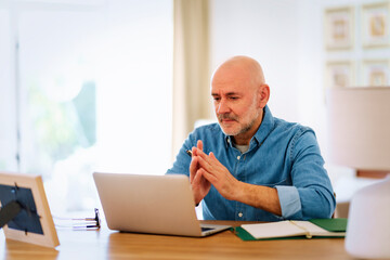 Confident middle-aged man sitting at desk at home and using notebook