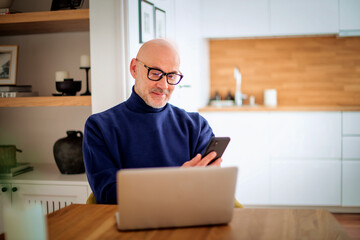 Confident middle-aged man sitting at desk at home and using notebook and smartphone
