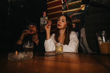 A small group relaxes in a dimly lit bar, with a woman smoking a cigar and drinks on the table, creating a warm, sociable nightlife mood.
