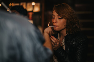 A woman wearing a leather jacket lights a cigarette in a dim, intimate night scene. The flame...