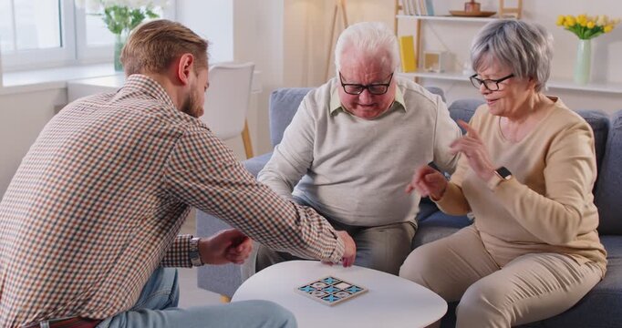 Happy senior and young people play tic tac toe on table in living room at home. Elderly father, mother and son with beard playing fun board game with wooden blocks, smiling and sitting on couch