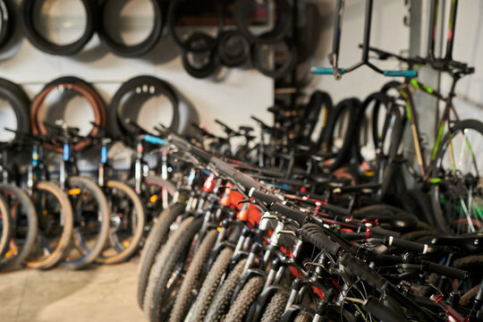 Row of mountain bikes standing in bicycle shop with multiple spare tires hanging on wall in background, showing organized display of cycling equipment for retail or rental