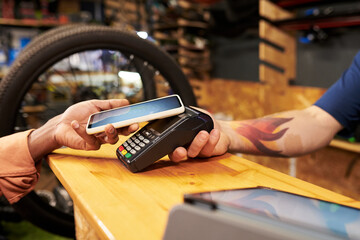Young man paying with smartphone using contactless payment terminal at bike shop counter, male employee with tattooed arm holding card reader during transaction