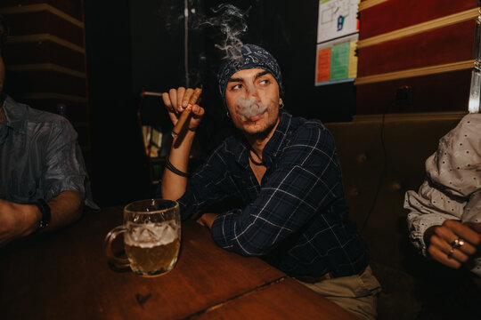 A man in a plaid shirt and bandana sits at a wooden bar, smoking a cigar as steam rises. A beer mug rests on the table, conveying a casual, nighttime social moment.