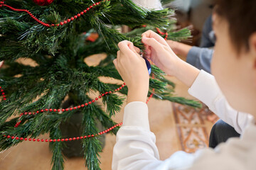 Unrecognizable toddler decorates a plastic Christmas tree with a red bead chain and hanging ornaments