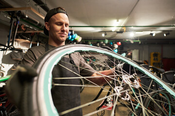 Young man wearing cap repairing bicycle wheel in workshop, holding bike rim with both hands, focused on maintenance task, surrounded by cycling equipment and tools