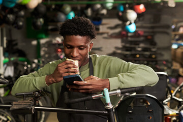 Young Black man standing in bicycle workshop eating sandwich while looking at smartphone, holding food in one hand and phone in other, surrounded by hanging helmets and bikes