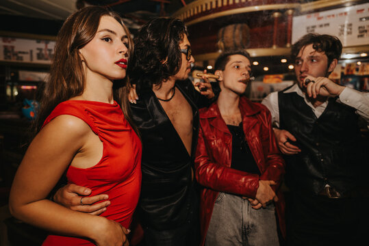 A stylish group of friends enjoys a night out at a bustling bar. A woman in a red dress stands close as others chat and smoke, capturing a lively urban party moment.