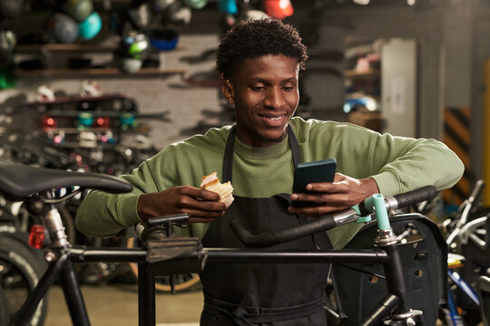 Young Black man wearing apron standing in bicycle workshop holding sandwich and smartphone, smiling while checking phone and leaning on bike repair stand with helmets in background
