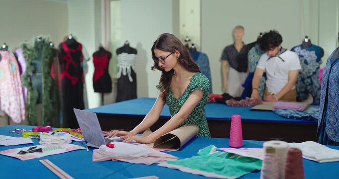 Indian young adult gen z female lady wear tape around neck learn clothe craft skill indoor class room. girl use laptop look screen typing hold pencil do fabric pattern on paper new day college place