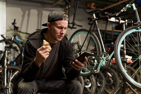 Young man wearing cap sitting in bicycle workshop eating sandwich while holding smartphone, surrounded by multiple bicycles and bike parts in background