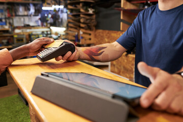 Young man with tattooed arm holding payment terminal while Black middle aged man making contactless payment with credit card at counter in retail store