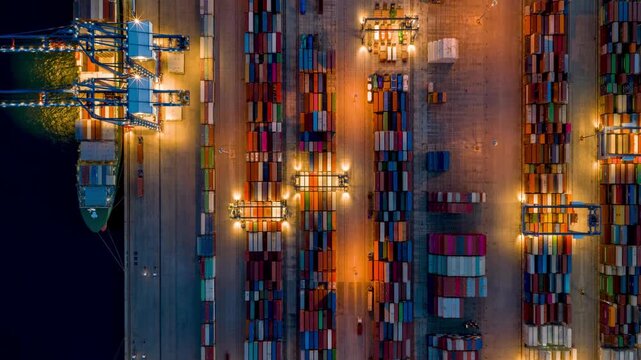 Aerial overhead motion time lapse view of a illuminated container terminal dock with loading and unloading activity by cranes and trucks off cargo ships during evening