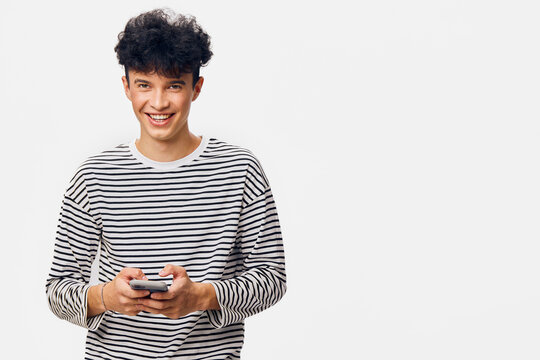 smiling man holding smartphone wearing striped shirt standing against white background with curly hair, casual style, youthful and positive expression