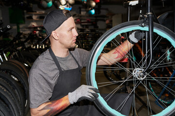 Young man wearing gloves repairing bicycle wheel in workshop, focusing on adjusting spokes, visible tattoos on arms, surrounded by multiple bikes in background