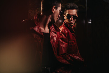 A stylish man in a red leather jacket and sunglasses stands beside a reflective surface in a low-lit setting, conveying attitude, mystery, and urban cool for fashion and lifestyle stock uses. © qunica.com