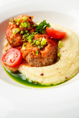 A gourmet dish featuring tender meatballs covered in rich tomato sauce, served alongside creamy mashed potatoes and garnished with fresh scallions and cherry tomatoes on a white plate.