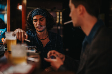 Two men sit at a bar, one with a headband smoking while the other hands him a cigarette, capturing a candid, moody nightlife moment.