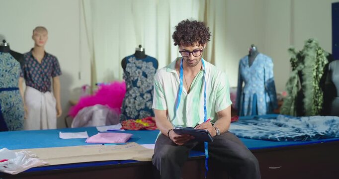 Indian young adult gen z smart male man hold tab learn new clothe craft course look camera class room. Boy sitting on desk table wear glass tape around neck staring cam do causal pose enjoy day place