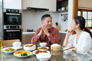 Middle aged Asian man and middle aged Asian woman sitting at kitchen table enjoying meal together, engaging in conversation, various dishes and bowls of food arranged in front of them
