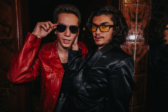Two men in bold outfits pose confidently at a doorway in a glossy, nightlife setting. One wears a red leather jacket, the other a black suit and tinted glasses, exuding stylish energy.