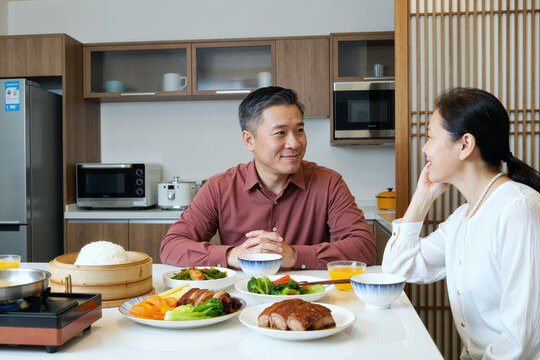 Middle aged Asian man sitting at dining table smiling and talking with middle aged Asian woman, sharing meal with various traditional dishes in modern kitchen setting