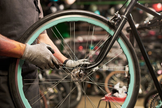 Young man wearing gloves repairing bicycle wheel in workshop, focusing on adjusting spokes and hub, hands visible holding tools, background showing multiple bikes - Powered by Adobe