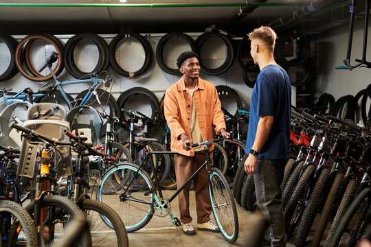 Young Black man holding bicycle while talking to young man in bike shop surrounded by various bicycles and tires, both standing and engaging in conversation
