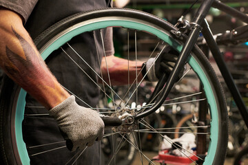 Young man with tattooed arm wearing gloves repairing bicycle wheel in workshop, focusing on tightening bolts and adjusting spokes, partial body visible