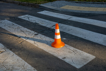 An orange traffic cone rests on a cracked crosswalk in an urban area, with sunlight casting shadows on the pavement