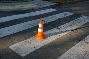 A bright orange traffic cone is positioned on a faded and cracked pedestrian crosswalk under sunlight, indicating a blocked path