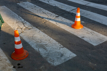 Two orange traffic cones are placed next to a pedestrian crosswalk on a cracked pavement as shadows stretch in the afternoon light