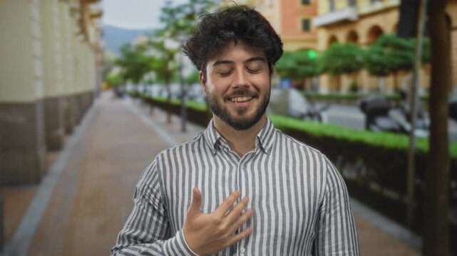 Young man with a beard standing outdoors on a street, wearing a striped shirt, smiling with a hand over his heart, surrounded by blurred greenery and buildings under a clear sky.
