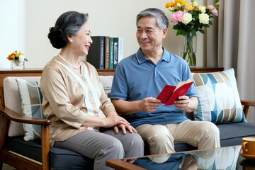 Senior Asian couple sitting on sofa smiling at each other, man holding red envelope, woman resting hands on lap, bookshelf and flowers in background