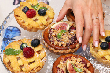 Woman hand reaching for fruit tartlets with berries, figs, and crumble topping. Homemade sweet dessert on glass plate. Delicious pastry with sugar powder and mint. Finger food background.