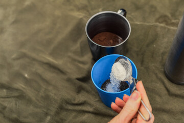 Young woman hiker making morning hot cocoa while sitting in camp tent. Adding powdered sugar to cup for making sweet hot cocoa outdoors