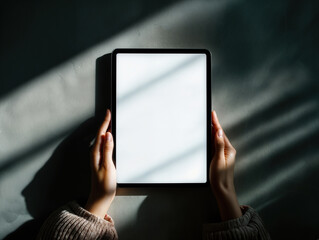 Tablet screen mockup held in hands with blank white display against dramatic light and shadow background. Clean digital template emphasizing modern technology, contrast, and copy space.