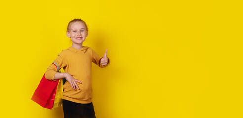 Smiling young girl holding colorful shopping bags and giving a thumbs up, standing against a bright yellow background.