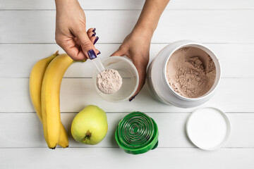Top view of female hand with measuring scoop putting chocolate whey protein powder into shaker on white wooden background, with protein jar, bananas and green apple, process of preparing protein drink