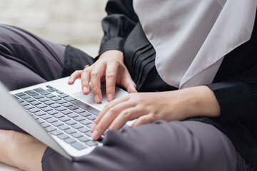 A close-up shot of A young Muslim woman's hand's in a grey hijab is sitting outside her house, focused on her laptop. Indonesian home terrace. A peaceful image representing modern Muslim lifestyle