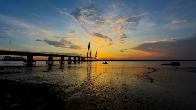 A dramatic sunrise/sunset silhouette of the Sungai Johor Bridge (Jambatan Sungai Johor), a key cable-stayed bridge on the Senai-Desaru Expressway (E22) in Johor, Malaysia, with colorful, reflective wa