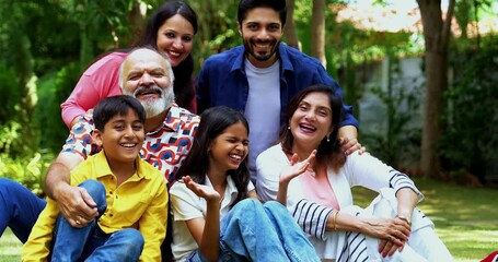 Indian family sitting on lawns smiling at camera, three generations including grandparents parents and children bonding together outdoors in park, happy Asian family enjoying leisure time together - Powered by Adobe