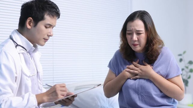 Medical professional providing attentive care, listening to female patient reporting breathing difficulties while documenting symptoms on clipboard within hospital room.