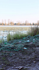  Photograph of an urban park wetland covered with plastic waste and greenery along the edges. Taken from ground level with frozen teal-colored fog, scattered grass patches, and clear blue sky above.